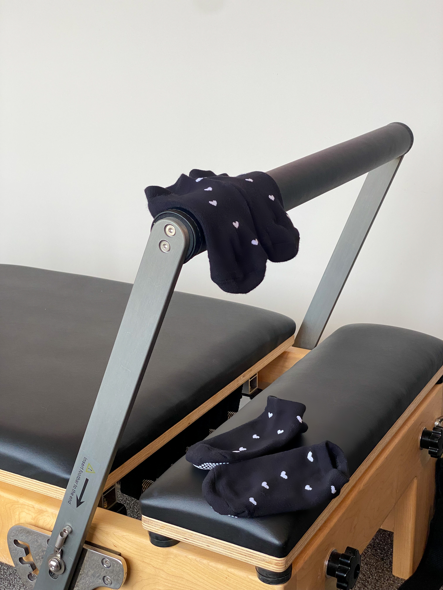 Black pair of Flow Peppy, New Zealand made ankle pilates and yoga grip socks with small white love hearts sitting on a reformer pilates bed.