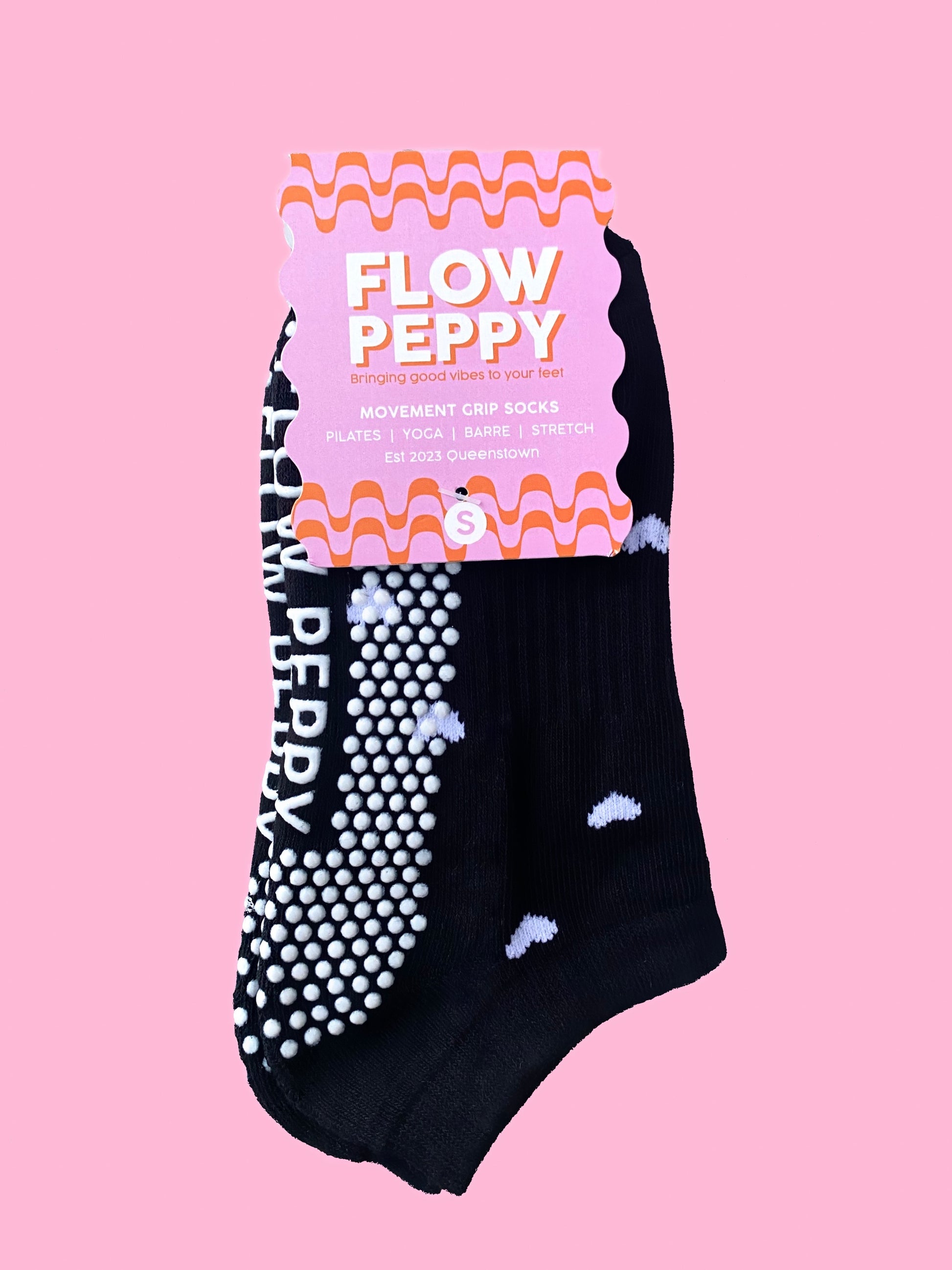 Black pair of Flow Peppy, New Zealand made ankle pilates and yoga grip socks with small white love hearts in packaging. 